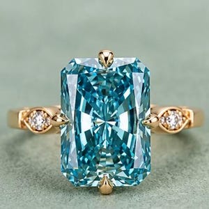 Könnte beinhalten: Ein Goldring mit einem großen, rechteckigen, leuchtend blauen Edelstein im Radiant-Schliff, der von vier Krappen gehalten wird. Die Ringschiene ist auf beiden Seiten mit einem kleinen, runden, klaren Edelstein in einer Goldfassung verziert.