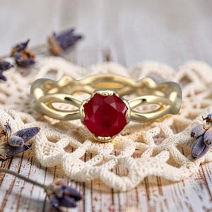 Könnte beinhalten: Ein goldener Ring mit einem runden, facettierten, tiefroten Edelstein in der Mitte. Die Ringschiene ist mit einem komplizierten, durchbrochenen, gedrehten Design gestaltet, das sich um den Finger legt und ein poliertes Finish sowie einen roten Stein zeigt.
