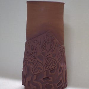 May include: A tall, cylindrical terracotta vase with a smooth upper section and a textured lower section featuring carved, geometric, and leaf-like patterns. The vessel has a deep reddish-brown matte finish and a flat, open top rim.