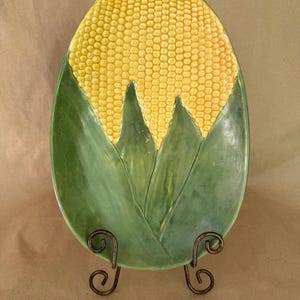 May include: An oval ceramic platter featuring a raised, textured yellow corn cob design at the top, transitioning into smooth, green, leaf-like shapes at the bottom. The platter is displayed on a dark brown, scrolled metal wire stand.