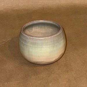 May include: A small, round ceramic bowl with a smooth, glazed finish. The exterior features a soft gradient of muted pastel colors, blending shades of pale green, light blue, and subtle pink tones. The rim is rounded and the interior is hollow.