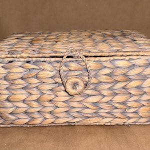 May include: A rectangular storage box crafted from woven light brown natural fibers. The box features a hinged lid and a front closure mechanism consisting of a braided loop that secures over a small, round, woven button-style fastener.