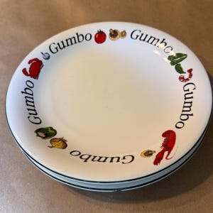May include: A white ceramic bowl with a black rim and the word Gumbo printed repeatedly around the edge. The rim features colorful illustrations of a tomato, mushrooms, green peppers, an onion, a crab, a shrimp, and a lobster.