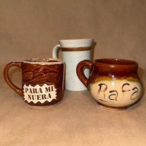 May include: Three ceramic mugs: one brown mug with a sombrero relief and the text "PARA MI NUERA", one tall speckled light blue mug with a brown band, and one rounded brown and cream mug with the handwritten name "Rafa" on the front.