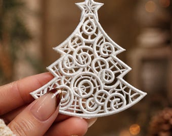 May include: A white embroidered Christmas tree ornament featuring an intricate openwork design of swirling circular patterns and a star at the top. The ornament is constructed from dense white thread with a small loop at the apex for hanging.