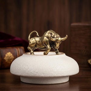 Può includere: Una statuetta di toro color ottone metallizzato posizionata su una base circolare in ceramica bianco sporco con finitura opaca e testurizzata. Il toro è raffigurato in posizione di carica con la testa abbassata, le corna in avanti e la coda alzata.