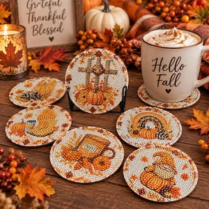 May include: A set of six round coasters featuring diamond art designs with autumn themes including pumpkins, a cross, a coffee mug, a rainbow, and a gnome. The coasters are decorated with small, reflective beads in orange, brown, and gold tones.