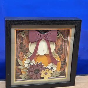 May include: A square shadow box frame with a black border containing a layered paper art piece. The design features a central white pumpkin topped with a dark red bow, surrounded by orange pumpkin segments, brown foliage, and various cream and purple flowers.