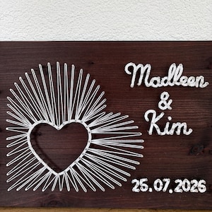 Könnte beinhalten: Ein dunkelbraunes Holzbrett mit weißer Fadenkunst in Herzform mit strahlenförmigen Linien. Der Text "Madleen & Kim" ist in weißem Faden über dem Datum "25.07.2026" angebracht, das ebenfalls aus weißem Faden besteht.