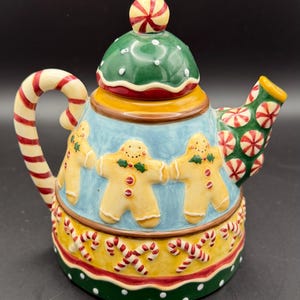 May include: A ceramic teapot featuring a light blue body decorated with gingerbread figures, a yellow base with red and white candy cane patterns, and a green lid topped with a peppermint candy knob. The handle is shaped like a candy cane.
