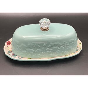 May include: A light blue ceramic butter dish with an oval base and a matching lid. The lid features an embossed floral pattern and a clear, faceted glass knob handle. The base has a scalloped edge with a colorful floral print pattern.