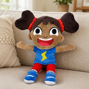 Puede incluir: Una muñeca de peluche con piel morena, cabello castaño oscuro en dos coletas con gomas rojas y ojos grandes. La muñeca viste una camiseta azul con un rayo amarillo, pantalones rojos y zapatos azules con suelas blancas, mostrando una amplia sonrisa.