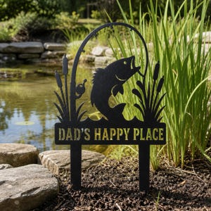 Puede incluir: Un estaca de jardín de metal negro con la silueta de un pez saltando del agua, una caña de pescar y juncos. El texto DAD'S HAPPY PLACE está recortado en una fuente negrita en la sección inferior, sostenido por dos puntas para el suelo.
