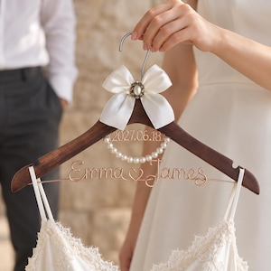 May include: A dark wood clothes hanger featuring a white satin bow with a central pearl-like embellishment at the hook. The hanger displays the date 2027.06.18, a circular string of white pearls, and the wire-script names Emma and James.