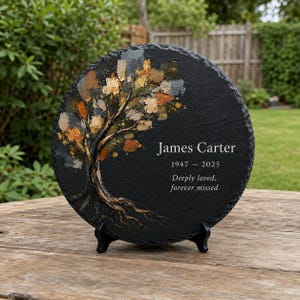 May include: A round black slate memorial plaque featuring a painted tree with textured gold, brown, and grey leaves. The text reads James Carter, 1947 - 2025, and Deeply loved, forever missed. The plaque is supported by a black plastic display stand.