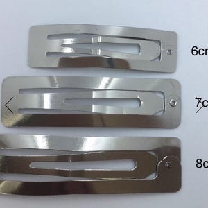 May include: Three silver-colored metal snap hair clips arranged vertically. The clips are labeled with their respective lengths of 6cm, 7cm, and 8cm. Each clip features a rectangular frame with a central spring-loaded mechanism for securing hair.