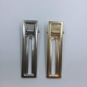 Peut inclure: Deux pinces à cheveux rectangulaires en métal, l'une de couleur argentée polie et l'autre de couleur dorée polie. Chaque pince présente un design à fente centrale ouverte et un mécanisme à ressort visible en haut pour maintenir les cheveux.
