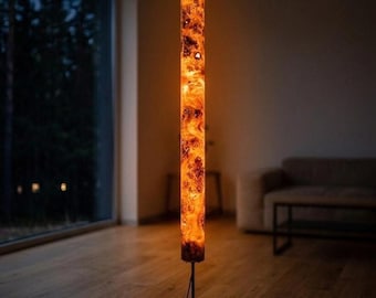 May include: A tall, cylindrical floor lamp featuring a glowing, amber-colored resin and wood burl body. The light fixture is supported by a thin, black metal pole and a square black base, emitting a warm, diffused orange light.