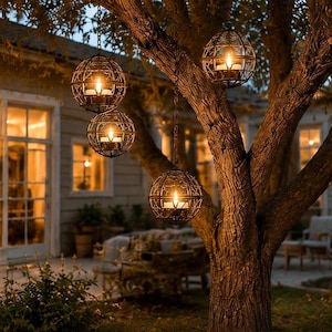 May include: Four spherical metal lanterns with a dark finish and intricate wire cage designs hang from tree branches by chains. Each lantern contains a glowing candle, casting warm light onto the surrounding tree bark and outdoor space.