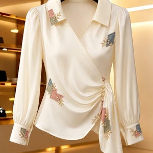 May include: A cream-colored long-sleeve blouse with a wrap-style front, featuring a pointed collar and gathered side detail with small white pearl-like buttons. The blouse is decorated with embroidered patches in muted pink and blue-grey tones.