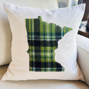 May include: A square off-white throw pillow featuring a centered applique in the shape of the state of Minnesota. The applique is made of a green, black, and white plaid fabric with a visible stitched edge outlining the state shape.