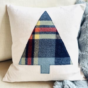 May include: A square off-white throw pillow featuring a centered applique of a Christmas tree. The tree is crafted from a blue, red, and yellow plaid fabric with a solid light blue trunk, all secured to the pillow with visible stitching.