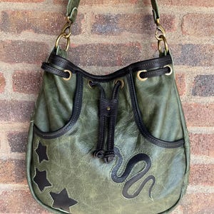 May include: An olive green leather hobo bag featuring a drawstring closure with dark brown leather trim. The bag is decorated with three dark brown star-shaped appliques on the left side and a dark brown snake-shaped applique on the front.