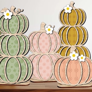 Puede incluir: Un conjunto de tres decoraciones de madera con forma de calabazas apiladas en tonos verde claro, rosa suave y amarillo mostaza. Cada calabaza tiene un patrón floral y una pequeña flor blanca con centro amarillo en la parte superior.