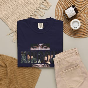 May include: A navy blue t-shirt featuring a graphic print of a person's eyes and profile with the text XINGOU overlaid across the center. The shirt has a crew neckline and a Comfort Colors brand label at the inner neck area, displayed flat.