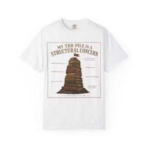 May include: A white t-shirt featuring a graphic of a tall stack of books with a small red flag on top. The text reads MY TBR PILE IS A STRUCTURAL CONCERN, with labels pointing to different books including Emotional Damage and Impulse Buys.