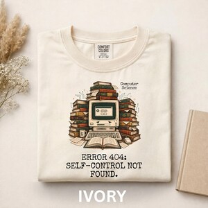 Könnte beinhalten: Ein elfenbeinfarbenes T-Shirt mit einer Grafik eines alten Computers, umgeben von Bücherstapeln. Der Text lautet "Computer Science" und "ERROR 404: SELF-CONTROL NOT FOUND." Das Design umfasst eine kleine Tasse, eine Maus und ein offenes Buch.