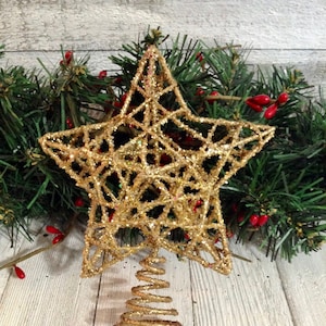 May include: A gold glitter star tree topper with a spiral base. The star is made of a wire frame covered in gold glitter. The background includes green garland with red berries, set on a white wooden surface.