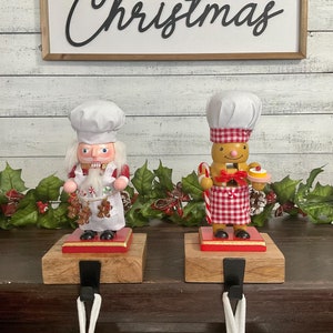 May include: Two wooden Christmas stocking holders featuring a gingerbread man and Santa Claus nutcracker. The gingerbread man is wearing a red and white checkered apron and holding a candy cane and a plate of cookies. The Santa Claus nutcracker is wearing a white chef's hat and apron and holding gingerbread cookies. Both are standing on wooden bases with black metal hooks.