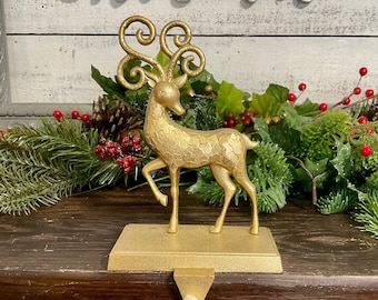 May include: Gold reindeer stocking holder with a white fabric loop. The reindeer has curly antlers and a textured surface. The holder is sitting on a wooden surface.