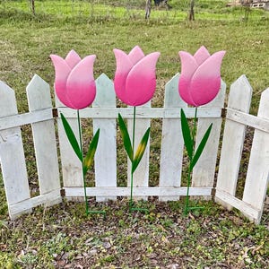 May include: Three pink metal tulips with green stems and leaves are displayed in front of a white picket fence. The tulips are painted with a gradient of pink, fading from a darker pink at the top to a lighter pink at the bottom.