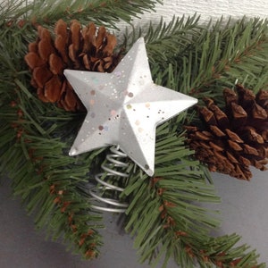 May include: A silver star Christmas tree topper with glitter accents, atop a silver metal spring. The star is surrounded by green pine needles and two brown pine cones, creating a festive holiday arrangement.