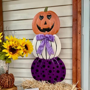 May include: A vertical metal pumpkin stack decoration featuring three pumpkins. The top orange pumpkin has heart-shaped eyes and a black smile. The middle white pumpkin has a purple and white gingham bow. The bottom purple pumpkin has black polka dots.