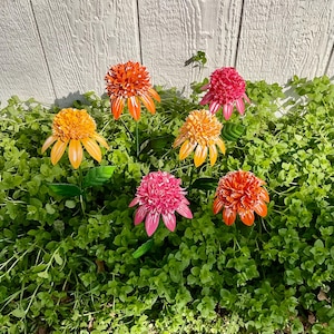 May include: Six colorful metal flower garden stakes. The flowers have orange, yellow, and pink petals with green stems and leaves. The flowers are set in a bed of green foliage against a white wooden fence.