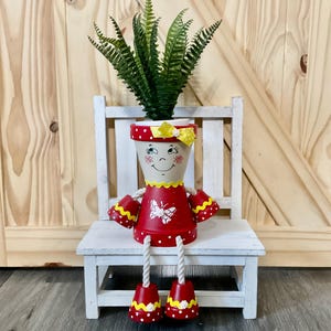 May include: A whimsical, hand-painted terracotta flower pot shaped like a doll, sitting on a small white chair. The doll pot has a red dress with a butterfly design, yellow polka-dot accents, and a smiling face. The pot holds a green fern.
