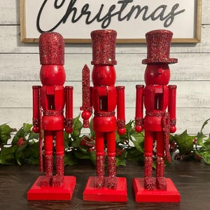 May include: Three red wooden nutcrackers with glitter accents on their hats and boots. One nutcracker holds a sword. A sign in the background reads "Merry Christmas".