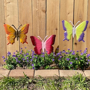 May include: Three colorful butterfly garden stakes. The butterflies are in shades of orange, red, pink, yellow, and purple. They are placed in a garden bed with purple flowers and a wooden fence background.