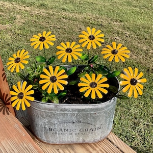 May include: An oval galvanized metal tub with the text "ORGANIC GRAIN PREMIUM QUALITY" printed on the front. The tub contains several yellow metal flowers with dark brown centers and green leaves, arranged to resemble a bouquet of black-eyed Susans.