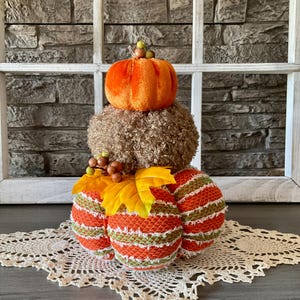May include: A decorative fall-themed arrangement featuring a stack of three pumpkin-shaped objects. The top pumpkin is orange velvet, the middle is brown, and the bottom consists of three orange and green striped pumpkins. Yellow leaves and berries add detail.