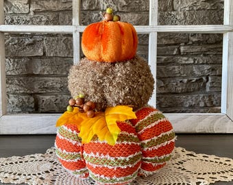 May include: A decorative fall-themed arrangement featuring a stack of three pumpkin-shaped objects. The top pumpkin is orange velvet, the middle is brown, and the bottom consists of three orange and green striped pumpkins. Yellow leaves and berries add detail.