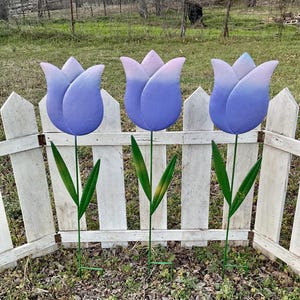 May include: Three metal tulip garden stakes. The tulip heads are painted in shades of blue and purple, with green stems and leaves. The stakes are positioned in front of a white picket fence, creating a decorative garden accent.