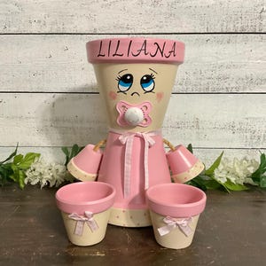 May include: A decorative terracotta pot craft shaped like a baby with the name LILIANA painted on the rim. The figure features a painted face with blue eyes, a pink pacifier, and a pink dress with a ribbon. Two smaller matching pots are in front.