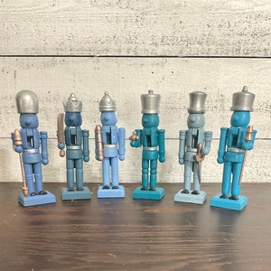 May include: Six blue and silver painted wooden nutcracker figurines standing on a wooden surface. Each nutcracker has a different hat and facial expression.