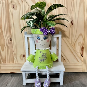 May include: A whimsical, hand-painted flower pot shaped like a seated figure, holding a plant with green and white striped leaves. The pot is lime green with white polka dots, a smiling face, and a purple bow. The figure is seated on a small white bench.