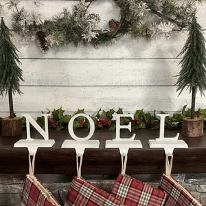 May include: White "NOEL" stocking holders with red and black plaid stockings hanging below. Two small artificial Christmas trees flank the letters. A garland of greenery and berries decorates the mantel, with a wreath above.