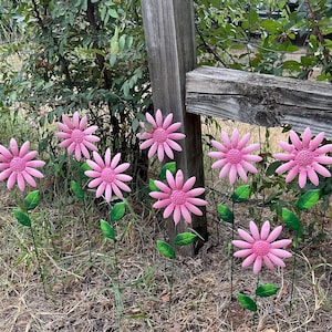 May include: Pink metal flower garden stakes with green leaves. The flowers have a detailed petal design and are arranged in a natural outdoor setting. The stakes are placed in the ground near a wooden fence.